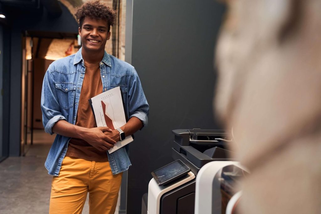 Ragazzo sorridente davanti ad una stampante in piedi con dei fogli in mano vestito con una camicia in jeans aperta, un t-shirt marrone e dei pantaloni color caki.