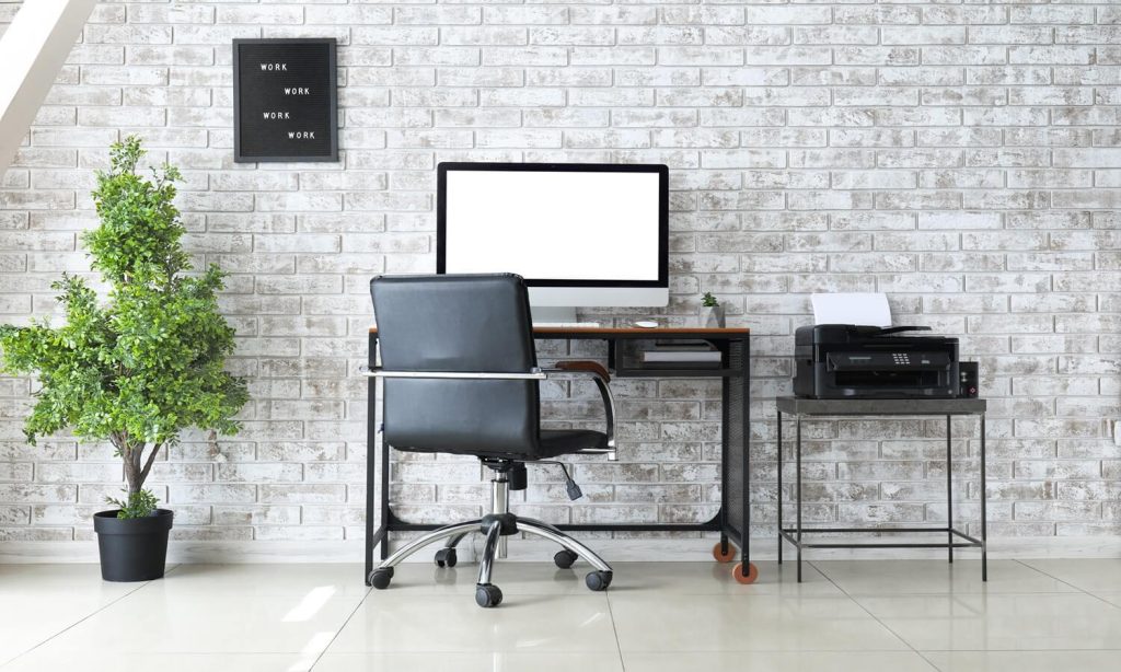 nell'immagine abbiamo un muro di mattoni bianco e davanti una scrivania, con un computer e una pianta verde sulla sinistra mentre a destra una stampante poggiata su un tavolino